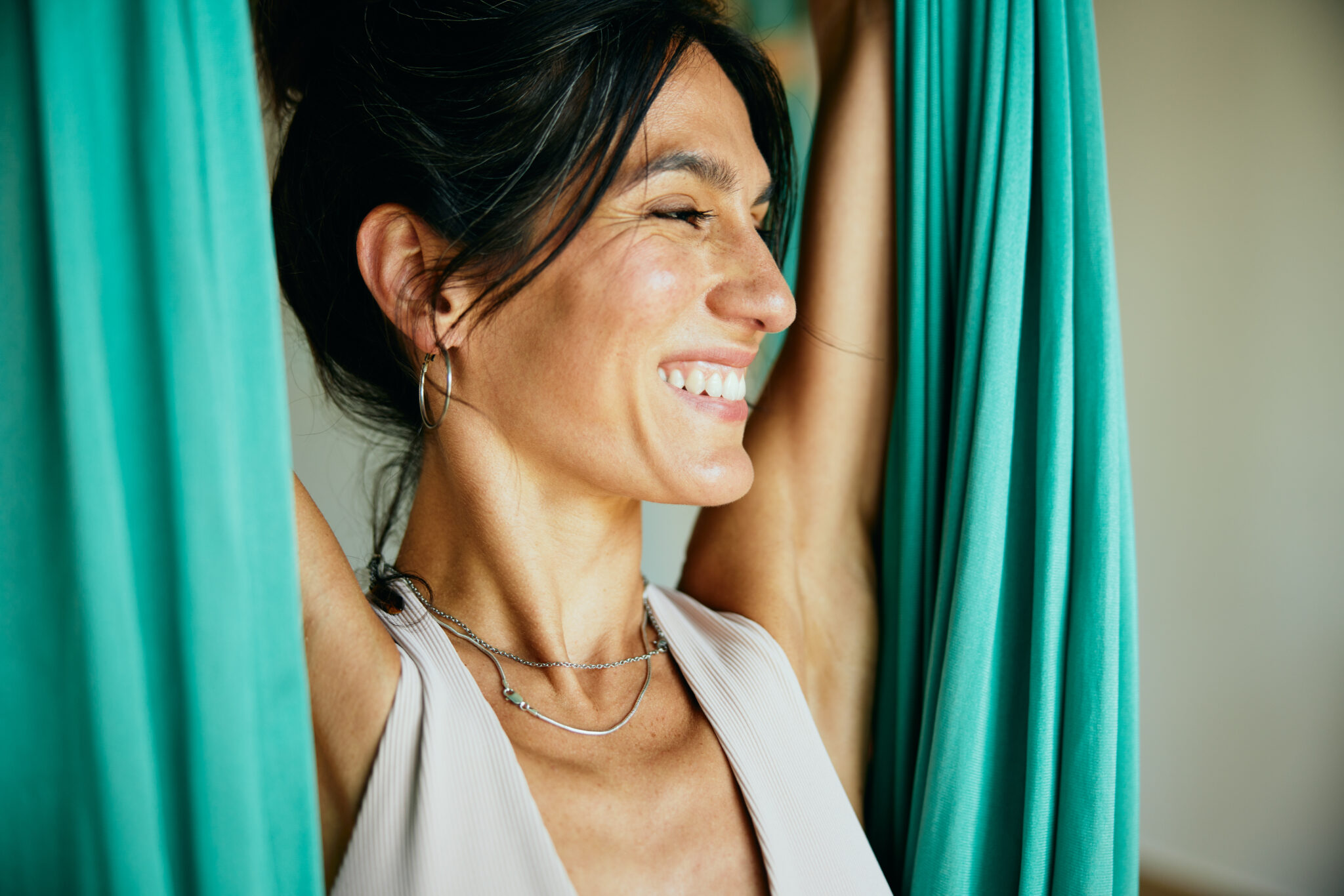 Fit woman in sportswear smiling while hanging from a hammock while practicing aerial yoga in a studio